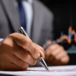 A person in a suit analyzing financial charts while writing notes with a pen