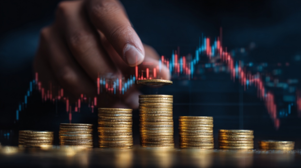 A hand placing a coin onto stacked coins with a fluctuating financial chart in the background