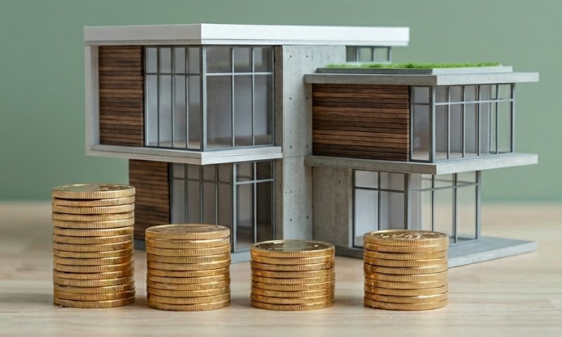 Model of a modern house with glass windows and wood accents, set beside stacks of gold coins